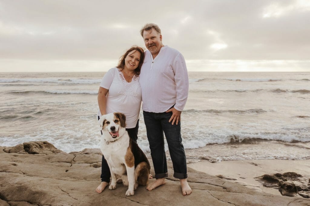 Couple and dog standing on rocks in front of ocean with overcast sky