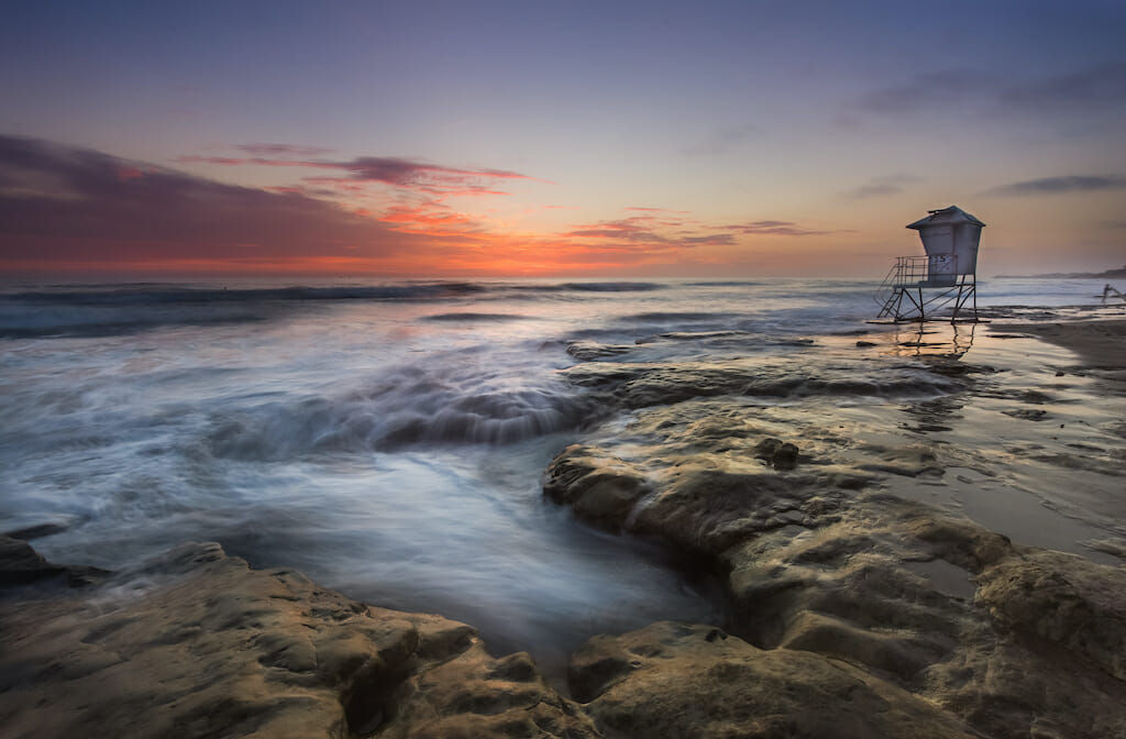 At sunset, the lifeguard stand at the Coronado beach is empty and the water is lively crashing against the natural terrain
