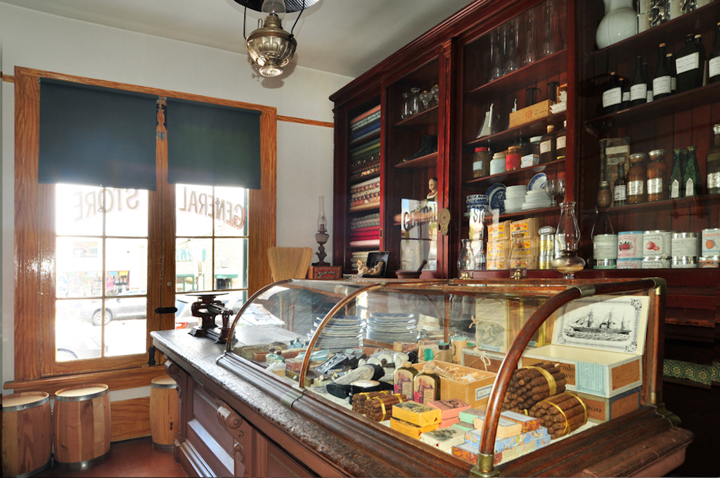 An old time general store with a wood and glass display case with knick knacks inside, the wall is lined with bottles and packages nd the sun shines through the window