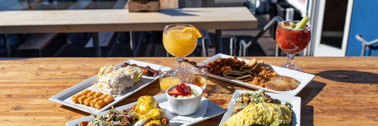 Several plates filled with breakfast foods near glasses of mimosas are displayed on a table with more chairs and the beach in the background