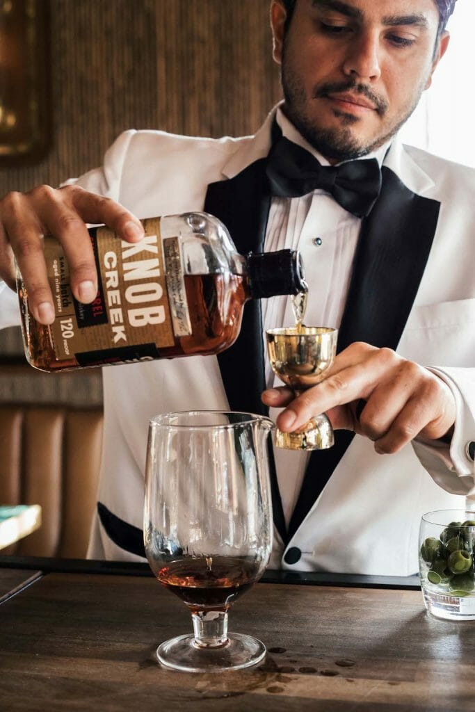 Bartender in white tuxedo pouring brown liquor into a jigger at the bar