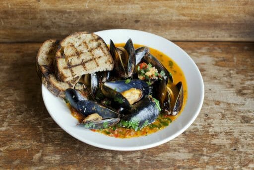 Two pieces of toast placed on the side of a bowl of mussels in a colorful broth