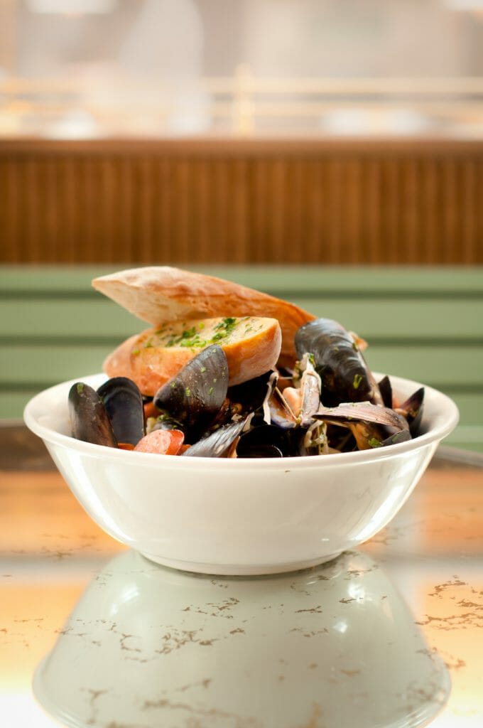 In a simple white bowl, beer braised mussels sit in their juices with two slices of crostini lay atop it