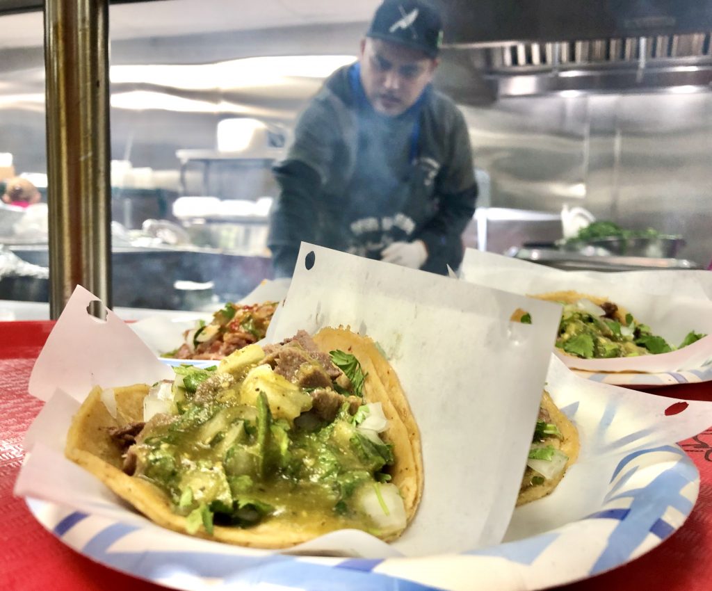 Plates with tacos on counter in front of man making the tacos in an open kitchen at Tacos El Gordo