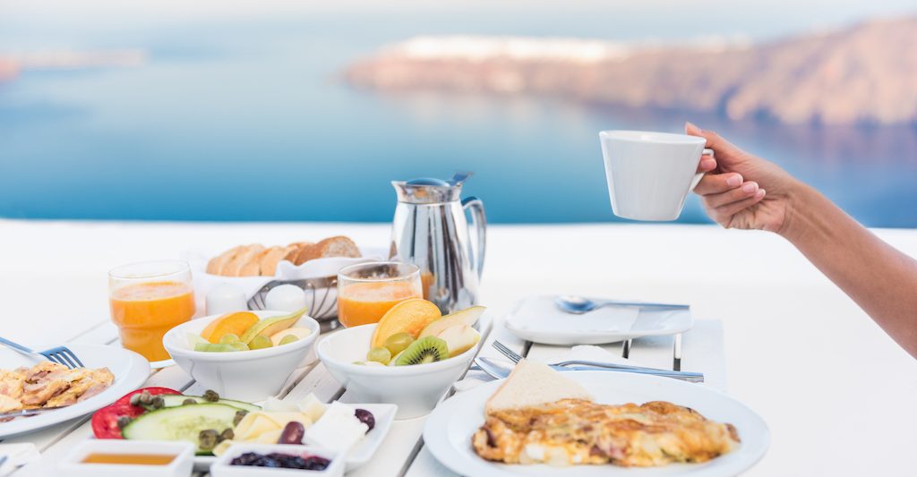 Morning person drinking coffee cup at breakfast table with mediterranean sea view. Woman eating at restaurant outside terrace patio on Santorini, Greece, Europe destination summer vacation.