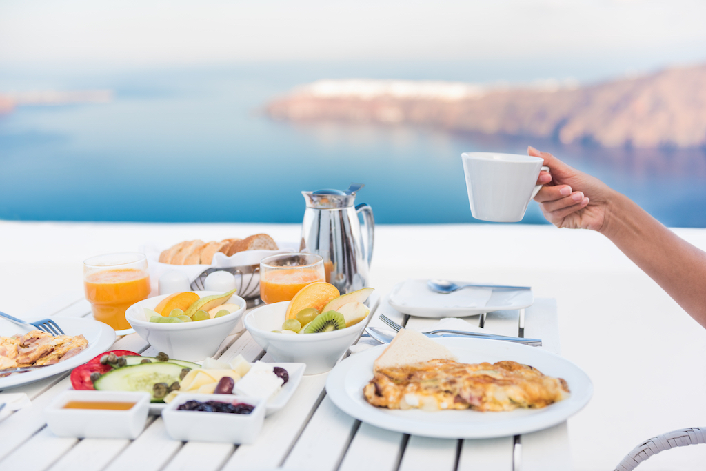 Morning person drinking coffee cup at breakfast table with mediterranean sea view. Woman eating at restaurant outside terrace patio on Santorini, Greece, Europe destination summer vacation.