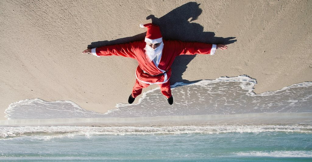 inverted image of santa claus lying face up on the sand of a beach, with the sea in the background