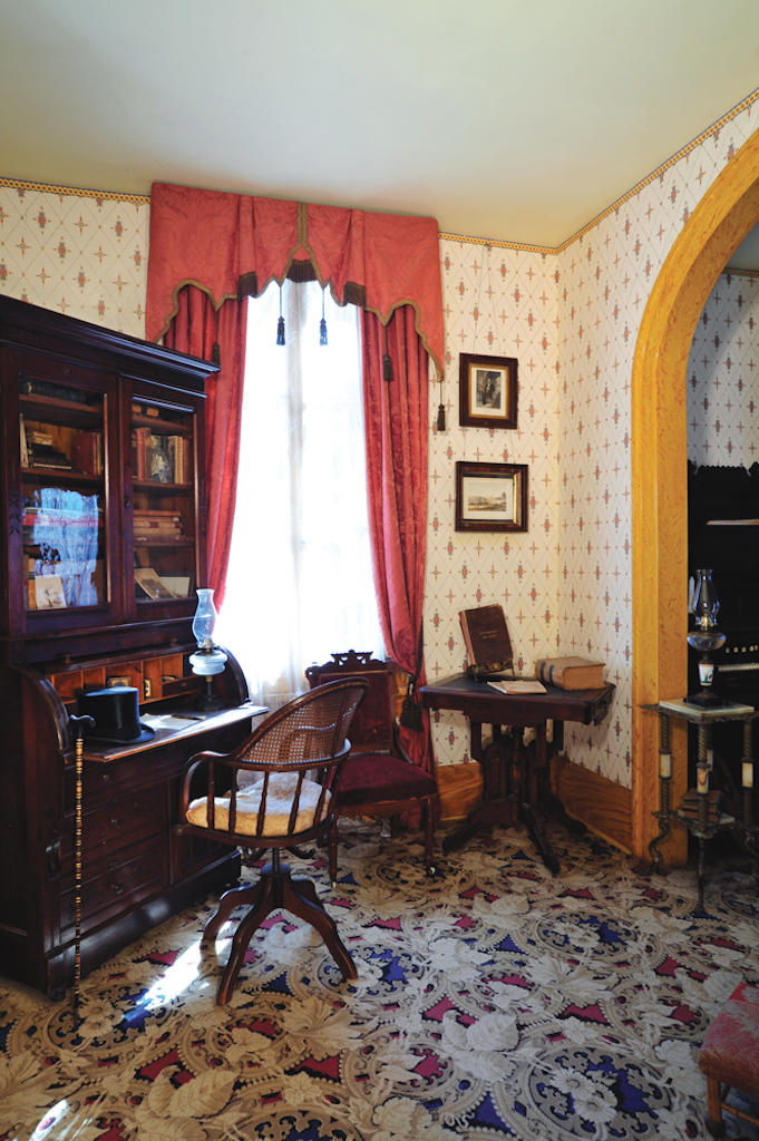 A study with cream and yellow wallpaper, windows covered with orange curtains and furnished with warm colored wood furniture