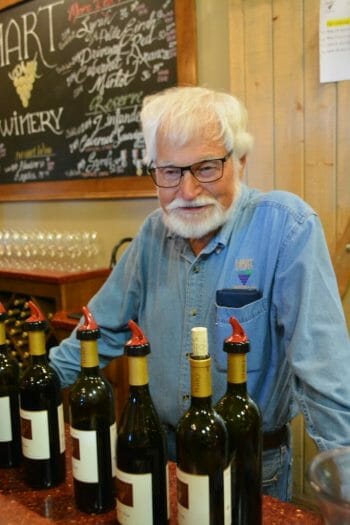 White haired man with beard, Joe Hart from Hart Family Winery Temecula, standing behind bar with wine bottles in front of him
