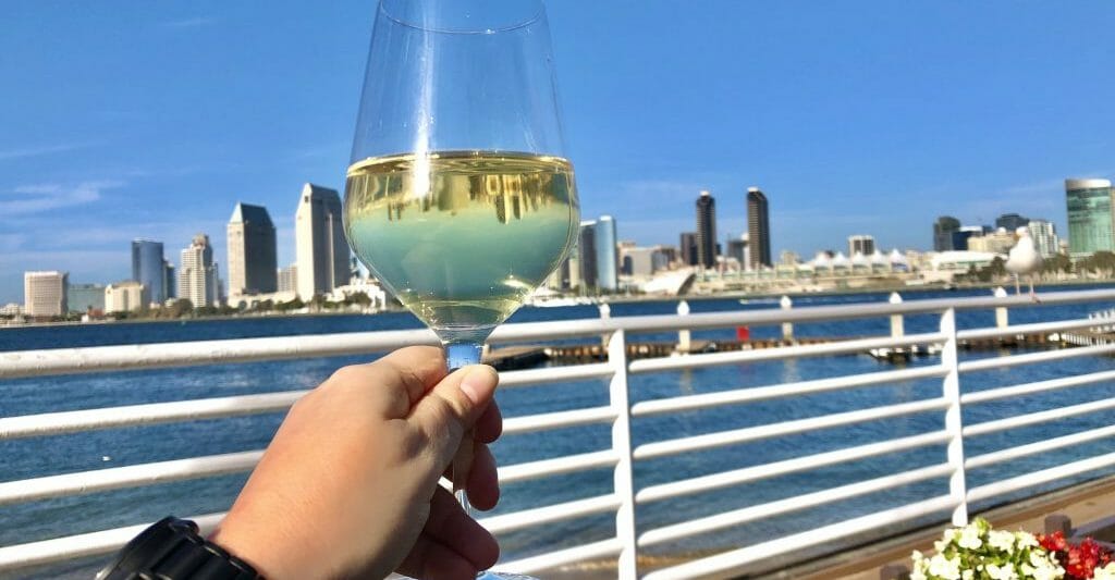 hand holding a wine glass with white wine with a white reeling and behind it the San Diego bay and the San Diego skyline
