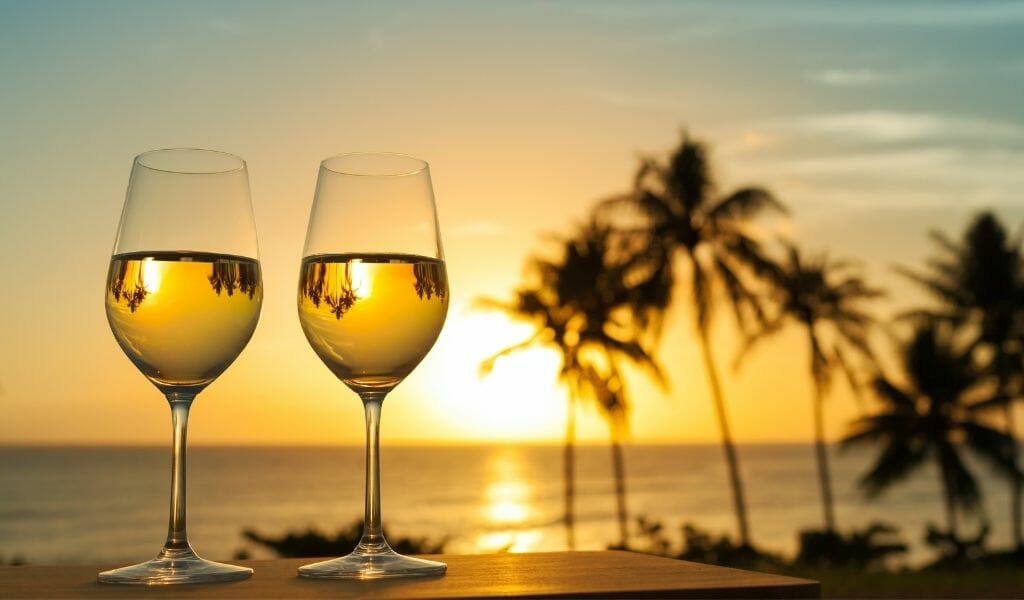 Two wine glasses  with white whine on a table overlooking the ocean at sunset with palm trees in the background