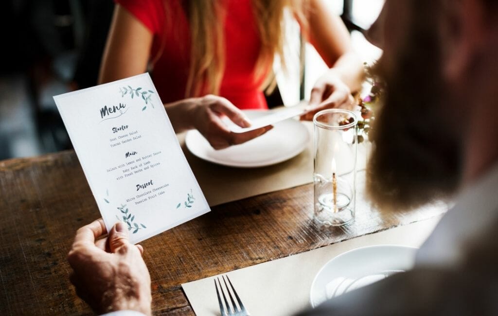Man holding a small paper menu while sitting at a restaurant table and a woman in a red top in the background blurry