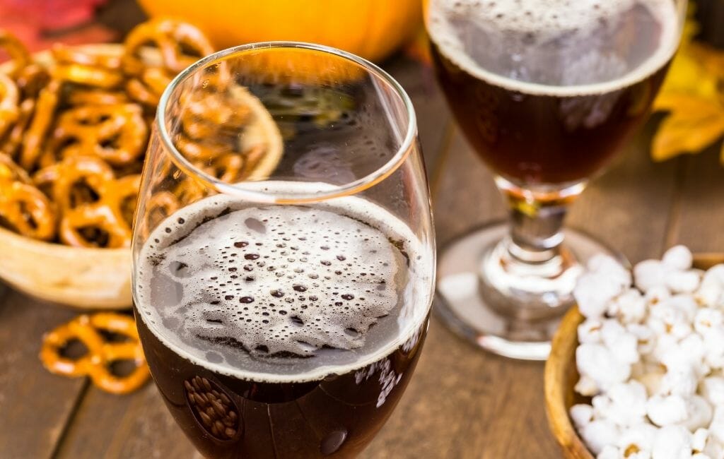 two glasses of dark beer on a table with a bowl of pretzels and pumpkins