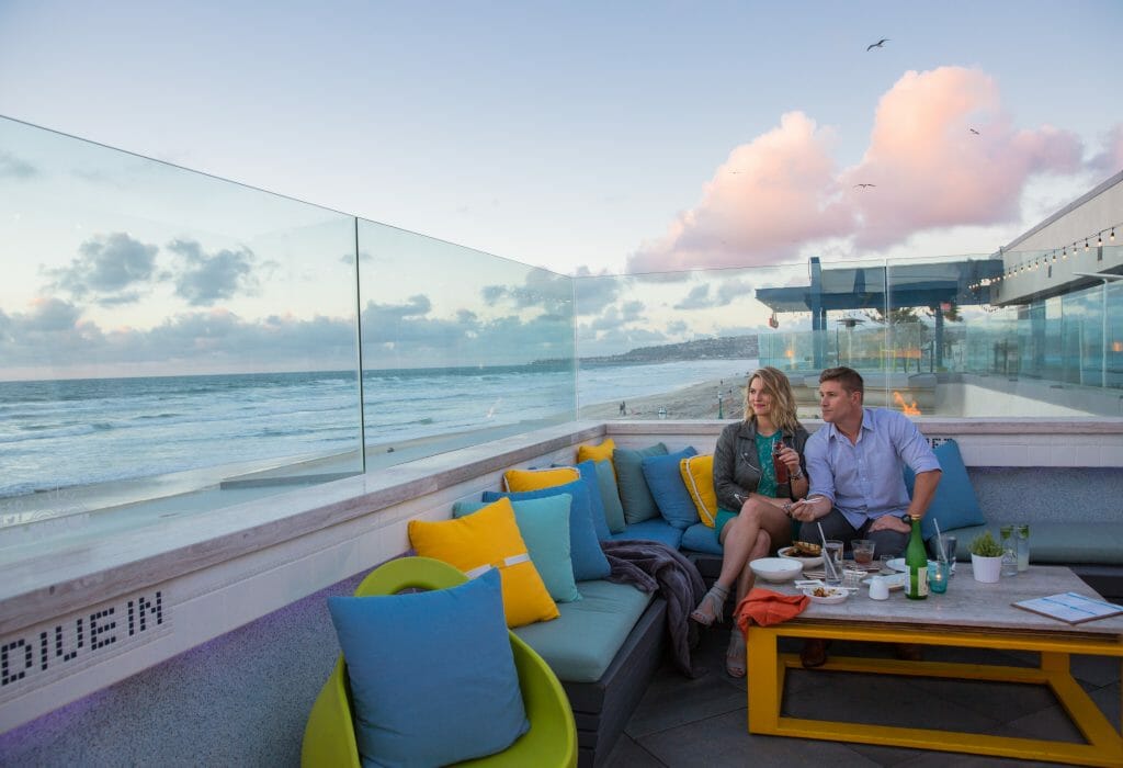 Couple sitting on a corner bench with blue and yellow pillows at Cannonball Rooftop bar looking out over the beach and ocean during sunset