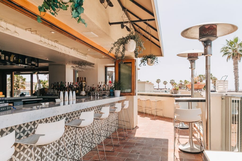 Outdoor Rooftop Bar At Catania La Jolla with a long white bar and white bar stools to the left and uncovered rooftop with heaters on the right
