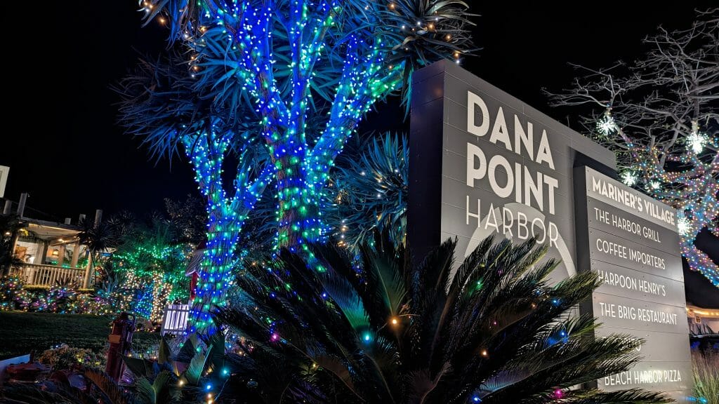 Sign with Text: Dana Point Harbor and list of restaurants and shops, trees decorated with blue and multicolored Lights