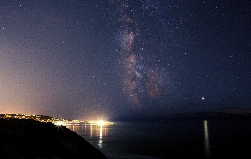 Milky Way photo with coastal cliffs on the left and ocean on the right, a lid up town on the horizon