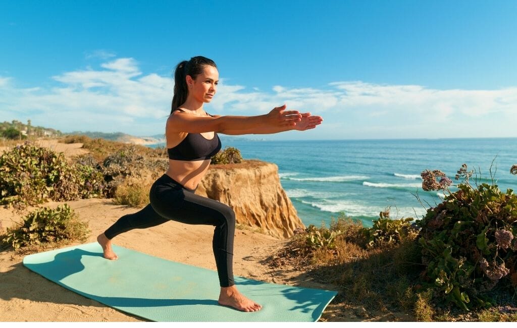Things To Do in Dana Point-woman doing yoga warrior pose on a turquoise yoga mat on a cliff overlooking the ocean 
