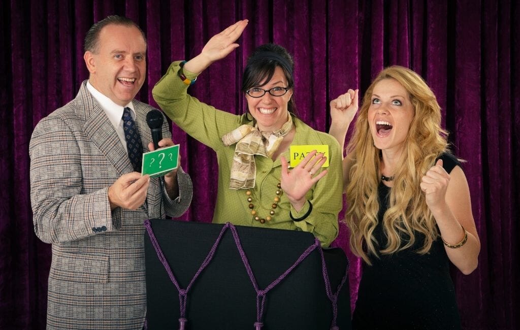 two women participating in a game show with male host in front of purple curtain