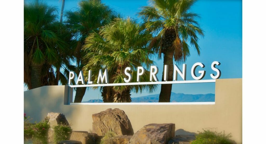 Wall with white metal sign "Palm Springs" in the foreground, palm trees and blue skies in the background