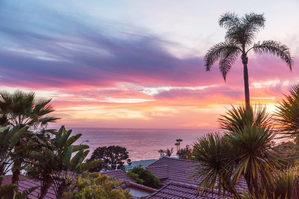 View overlooking spanish tiled roofs below with palm trees in-between, looking out over the Pacific ocean during stunning pink/purple sunset