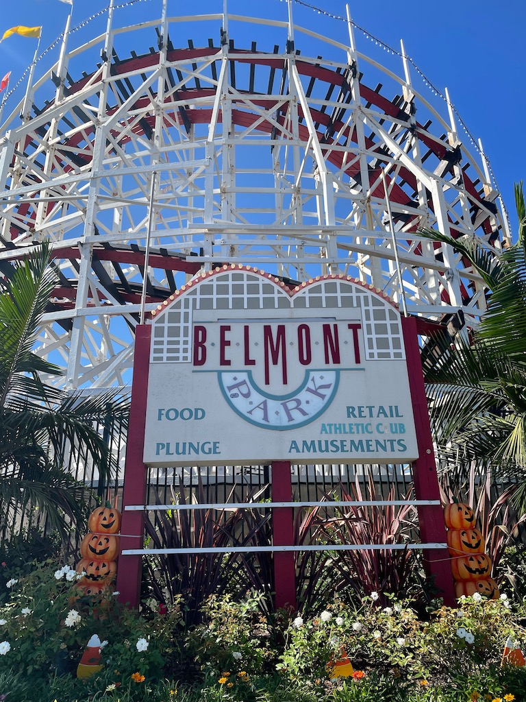 White and red wooden roller coaster with Belmont Park sign in front surrounded by green plants on a sunny day. Belmont Park Mission Beach roller coaster.