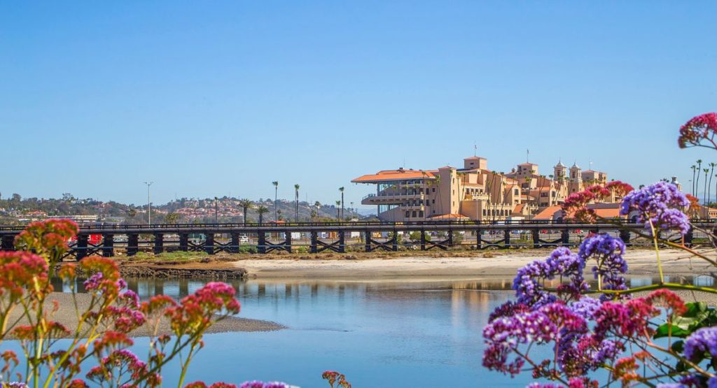 The large Del Mar Race Track building in distance with sandy shore and colorful flowers in foreground on sunny day. Del Mar, California. San Diego Coaster train stop.