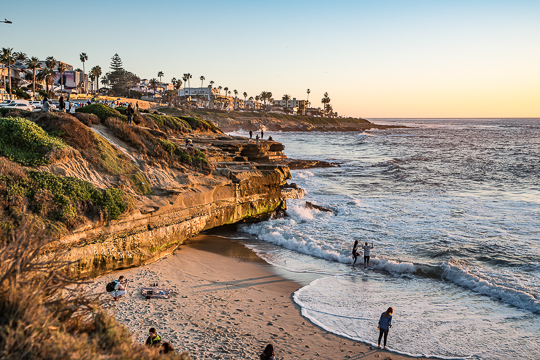 Sunset over sandy cove surrounded by rocky shoreline on Pacific Ocean, beach goers playing in water and searching tide pools. Shell Beach, San Diego tide pools.