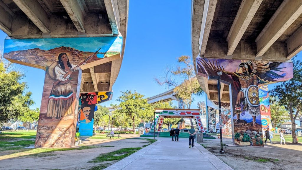 Large concrete beams painted in huge, colorful murals on highway underpass on sunny day. Chicano Park murals, Barrio Logan.