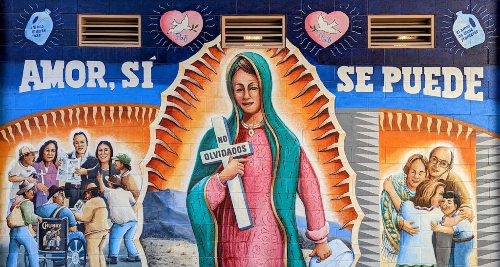 Large painted mural with the Virgin Mary holding a cross in the center surrounded by Mexican-American families, the mural reads "Amor, si se puede". Chicano Park murals.