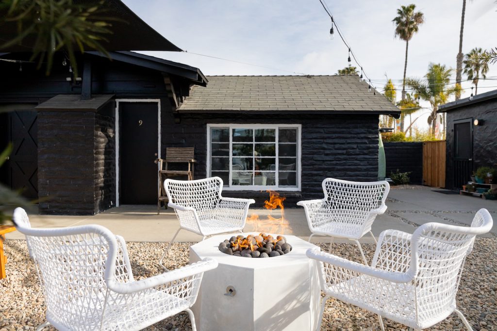 White wicker lounge chairs surrounding fire pit with small black building and palm trees in background. Green Room Hotel. Hotels in Oceanside California.
