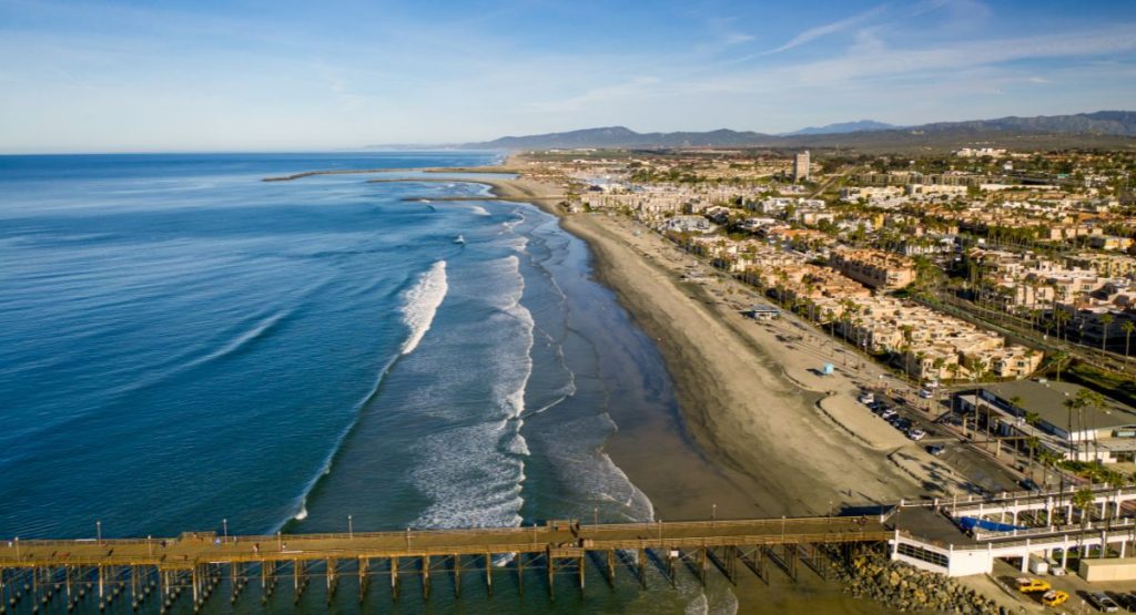 Ariel view of Pacific Ocean meeting sandy shoreline with long pier extending into water and low rise buildings overlooking beach, with mountains in distance on sunny day. San Diego Oceanside, California. Hotels in Oceanside