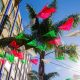 Red, green, and white paper flags with decorative cut outs, stringed above outdoor courtyard with building and palm tree in background on sunny day. Tijuana, Mexico Plaza Santa Cecelia. Tijuana tour from San Diego.