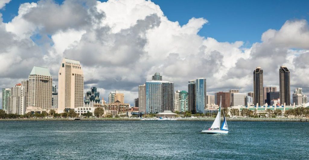 Small sailboat sailing calm waters with San Diego skyline in background on sunny day. Sailboat tours San Diego.