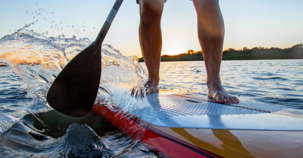 Close up action shot of man paddle boarding on calm water during sunset. Paddle board is colorful and water is splashing in the air has he paddles. SUP rentals in San Diego.