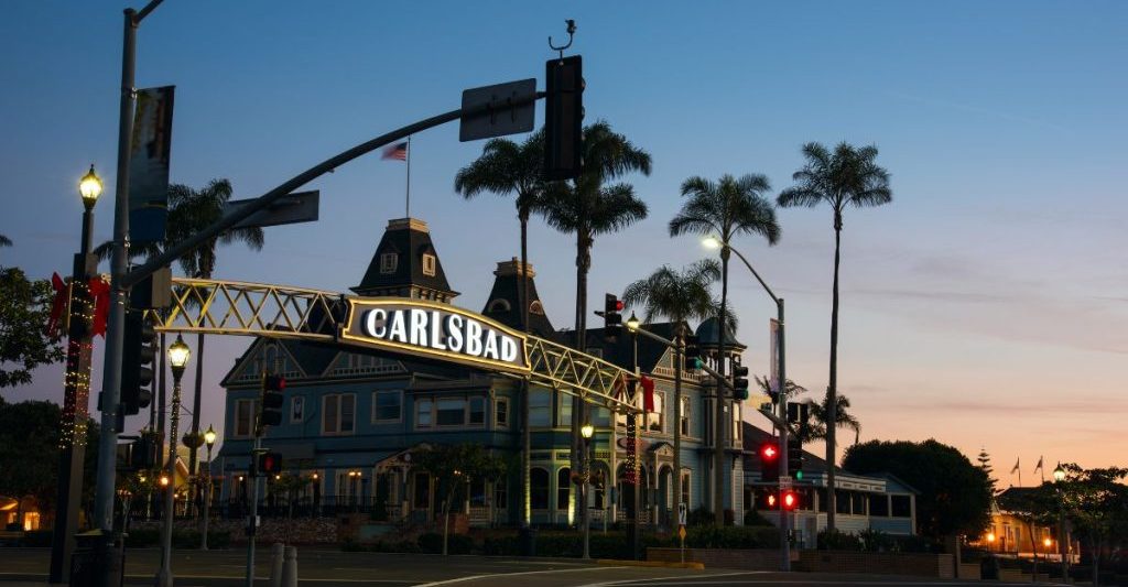 Large, backlit sign spanning street, reading "Carlsbad" in background are silhouettes of buildings and palm trees during late sunset. Carlsbad Village. What to do in Carlsbad.