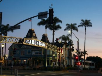 Large, backlit sign spanning street, reading "Carlsbad" in background are silhouettes of buildings and palm trees during late sunset. Carlsbad Village. What to do in Carlsbad.