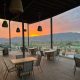 Wooden patio with restaurant table seating, patio is covered but completely open on sides with views of mountains in distance and desert valley below during bright orange sunset. Montevalle Health and Wellness Resort dining area.