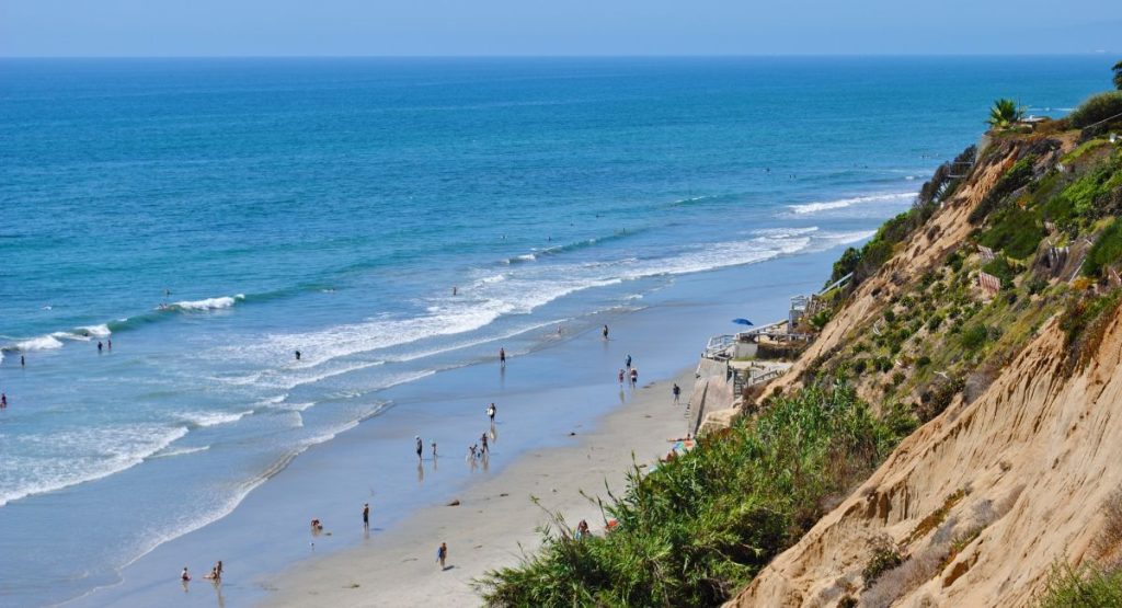 Sandy cliff covered in greenery against flat, white sand beach with beach-goers in Pacific Ocean on sunny day. Beaches in Carlsbad.