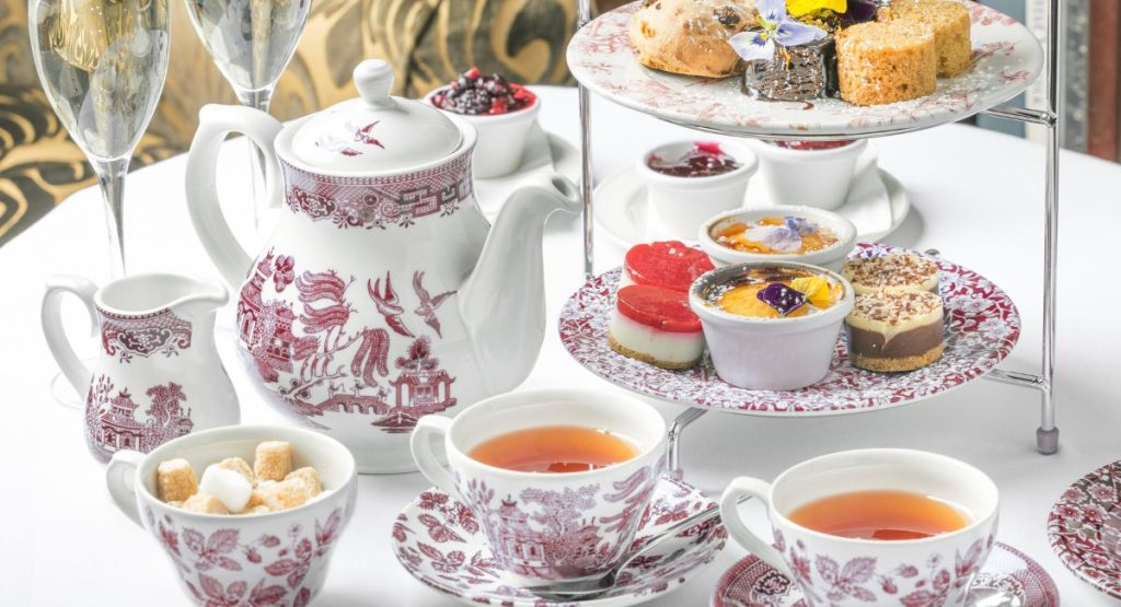 Fine China tea set with red florals and illustrations, matching tiered tray has finger foods and pastries on round table with white table cloth. High Tea San Diego.