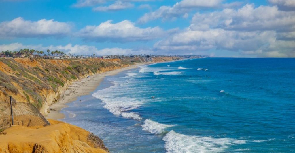 Blue water of the Pacific Ocean breaking on white sand beach next to sandy beach cliffs lined with palm trees on sunny day. North Ponto Beach, Carlsbad Beaches.