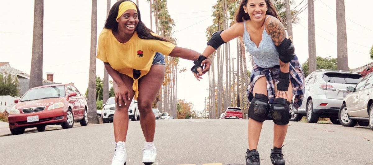 Two women holding hands while roller skating on street lined with cars and palm trees on hazy summer day. Roller Skating San Diego.