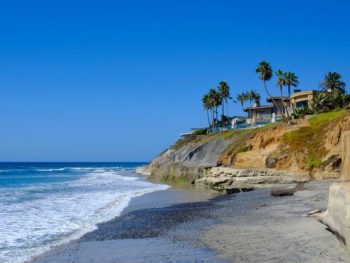 Waves breaking on sandy beach next to hill with large homes and palm trees on sunny summer day. Terramar Beach, Carlsbad Beaches.