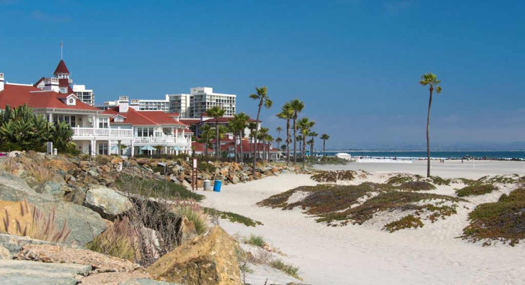 White sandy beach next to historic hotel with red roof surrounded by palm trees on sunny day with Pacific Ocean in background. Coronado Beach, San Diego in June