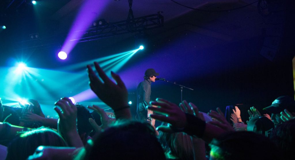 Man performing live music on stage in front of crowd of fans dancing to music, bright spot lights in background. Music Venues in San Diego.