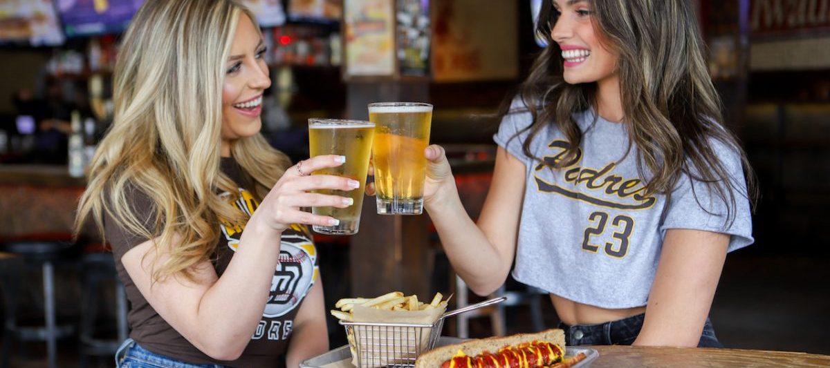 Two women in Padres gear at bar cheers-ing beers with plate of bar food. Best sports bars in San Diego, The Bootlegger.