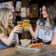 Two women in Padres gear at bar cheers-ing beers with plate of bar food. Best sports bars in San Diego, The Bootlegger.