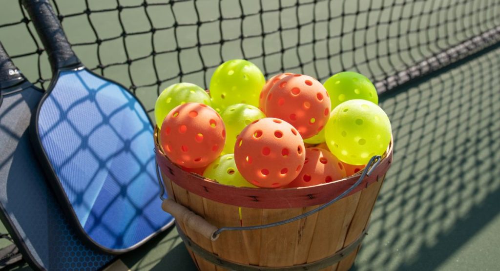 Bucket of orange and yellow pickleballs next to two blue pickleball paddles on outdoor pickleball court. Where to play Pickleball in San Diego.