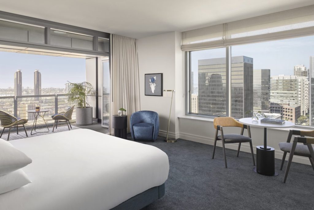 Hotel room with king size bed with white linens next to small seating area with large, open balcony overlooking city views on sunny day. Kimpton Alma, romantic getaways in San Diego.