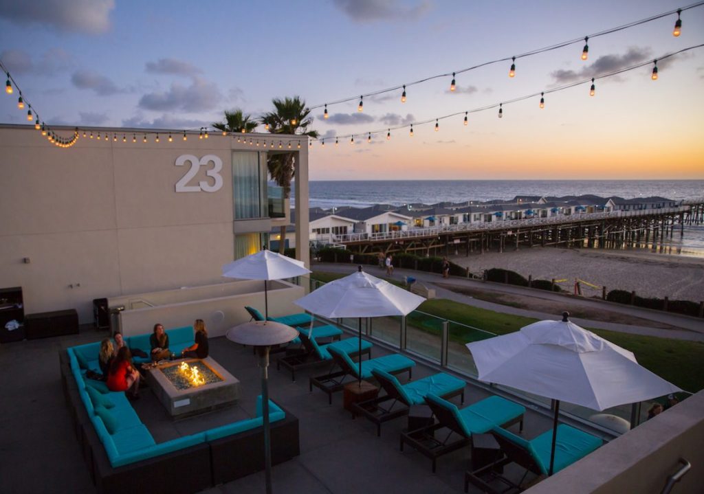Outdoor courtyard with colorful outdoor furniture, lounge chairs, umbrellas, and fire pit under string lights, overlooking boardwalk extending into ocean during sunset. Tower 23, San Diego beach resorts for couples.
